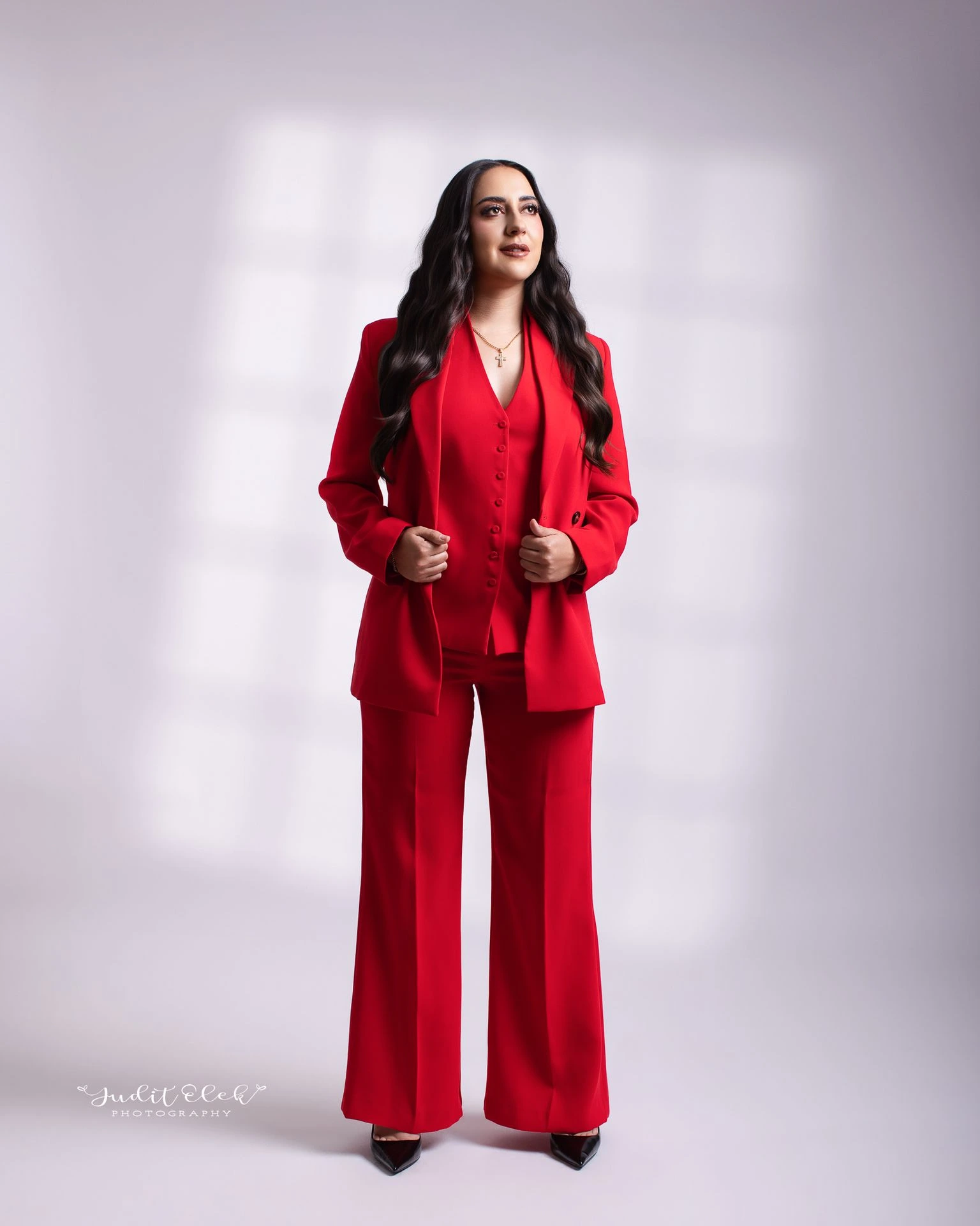 Retrato de cuerpo completo de mujer con traje rojo y manos sobre el saco, frente a un fondo gris claro.