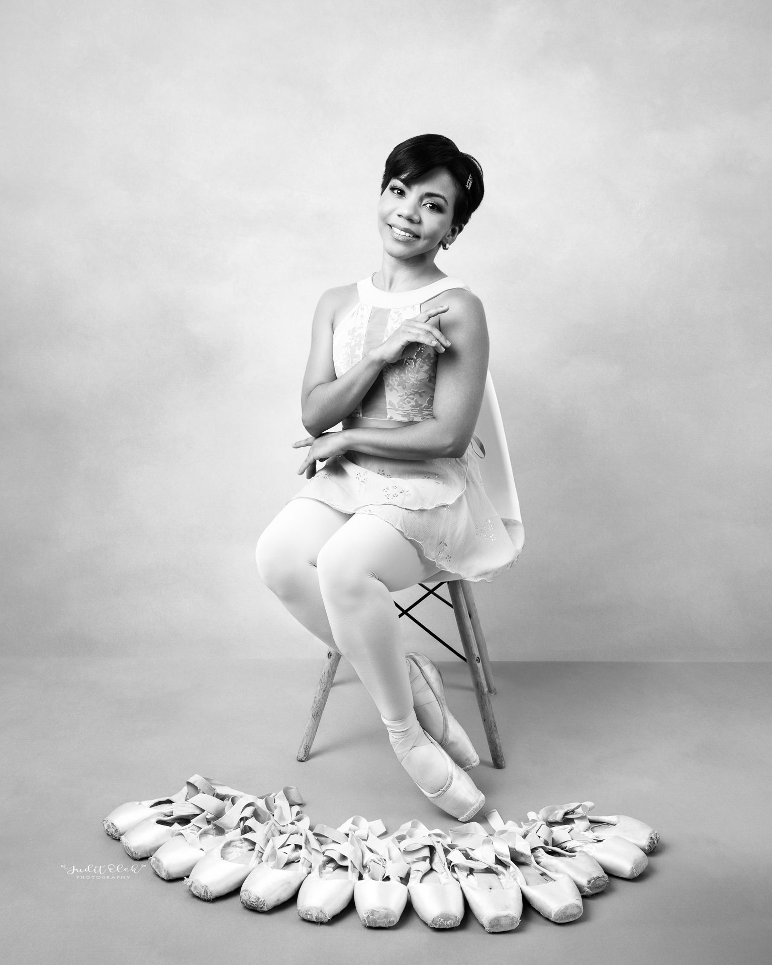 Fotografía en blanco y negro de bailarina sentada en un banco alto, con vestuario claro y pétalos sobre el suelo