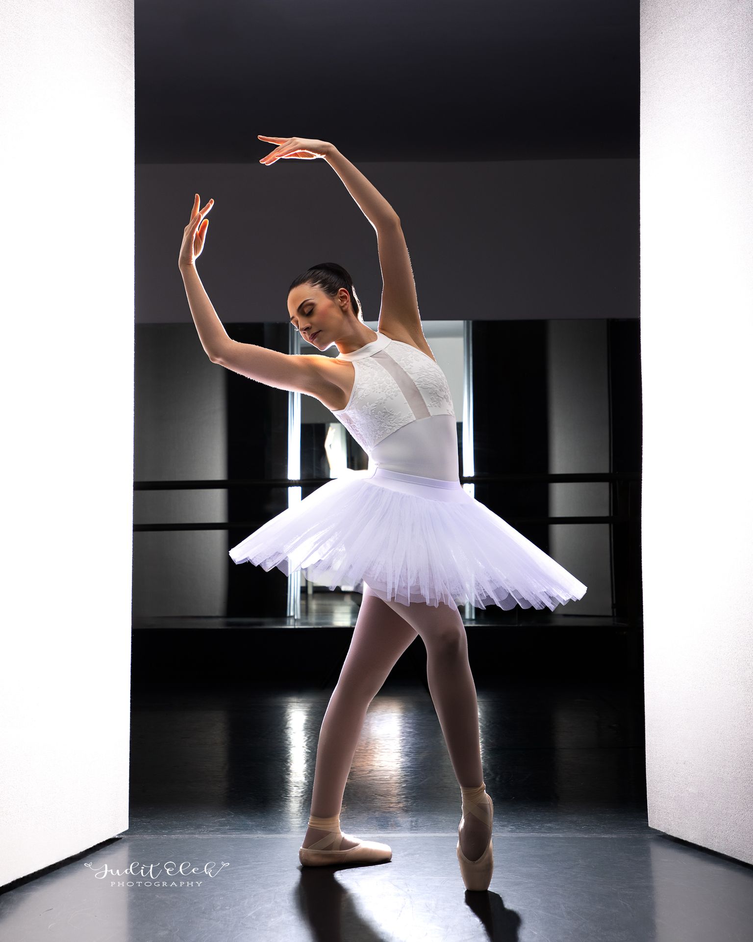 Bailarina con tutú blanco y zapatillas de punta, con los brazos elevados frente a un fondo oscuro.